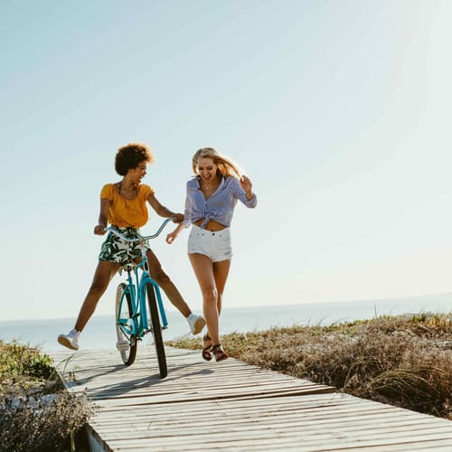 Two young women having fun with a bicycle at the beach. Woman running with friend riding a bike on boardwalk.